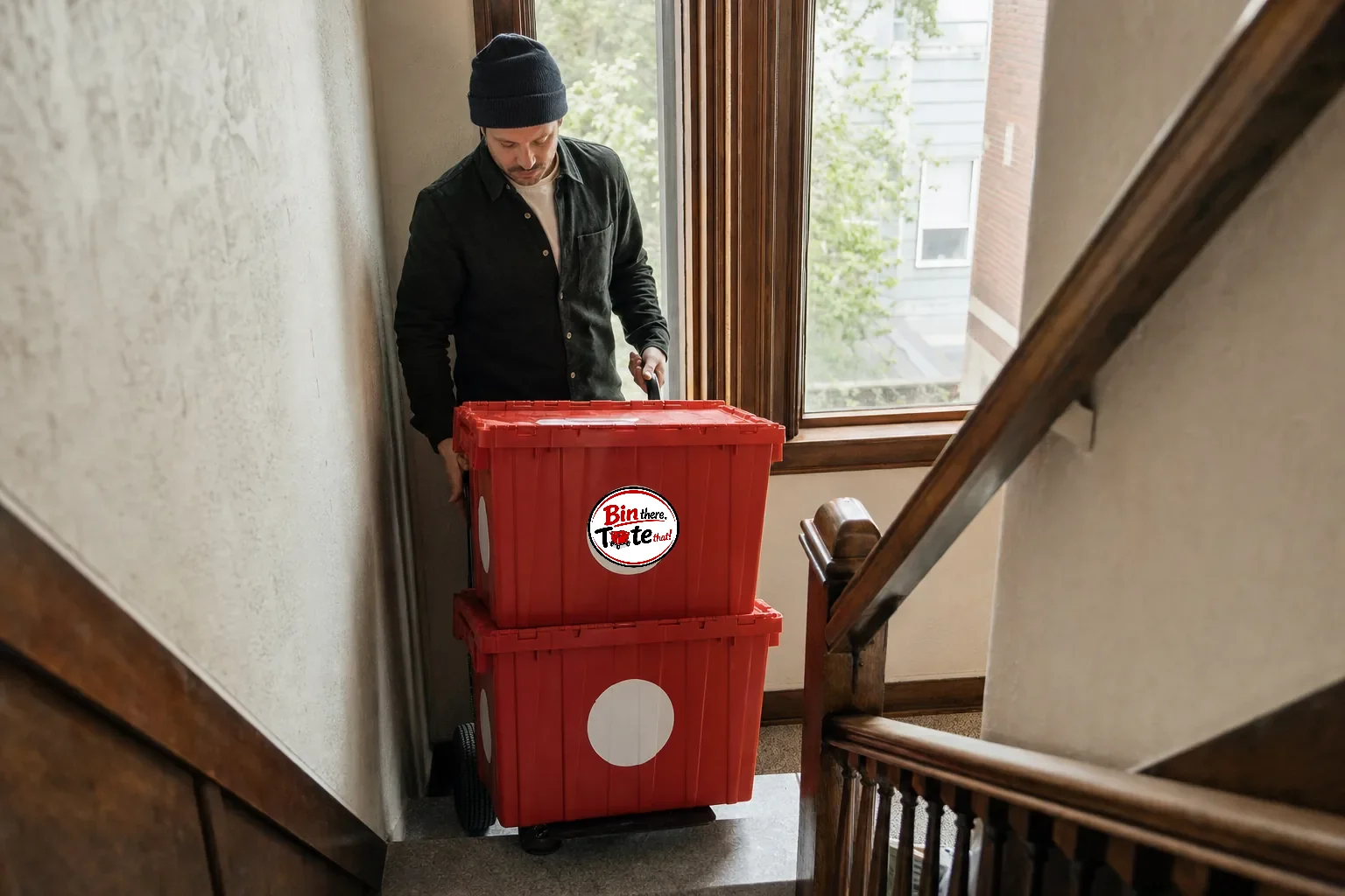 Person steadying a hand truck with two red moving totes on the landing of an older Portland apartment staircase